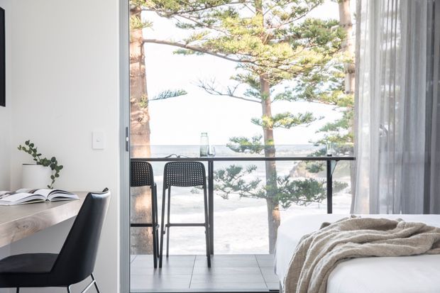 Bright, minimalist bedroom with a compact desk and black chair beside full-height glazing. The open glass door leads to a small balcony with two bar stools and a black railing, overlooking trees and a pale coastal or waterfront setting. A neatly made bed with a textured beige throw occupies the right side of the room.