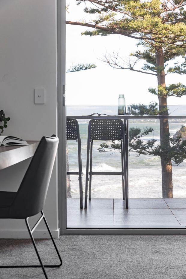 A bright contemporary dining area beside full-height sliding glazing, opening to a tiled balcony or terrace with two perforated bar stools and an ocean view. The interior has light grey carpet, white walls, a pale dining table, and a black modern dining chair. A sculptural coastal tree frames the outdoor view.