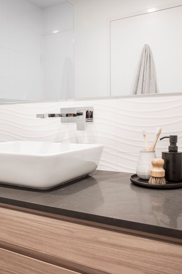Close-up of a contemporary bathroom vanity featuring a white vessel basin, wall-mounted chrome tapware, a dark grey stone benchtop, and light timber cabinetry. The backsplash has sculptural horizontal wave-textured white tiles, with a large mirror above and understated toiletries arranged on a black tray.