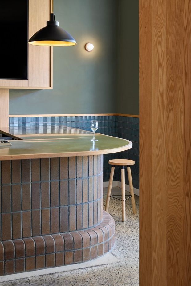 A close interior view of a compact bar or kitchen counter with a rounded peninsula clad in narrow dark terracotta and grey tiles, a pale green stone-like benchtop, and a curved tiled base. The space features dark blue-green vertically ribbed wall tiles, muted green painted walls, warm timber cabinetry and a timber door or screen, a black pendant light, and terrazzo flooring. A small timber stool and wine glass sit beside the counter, creating a refined retro-inspired hospitality atmosphere.