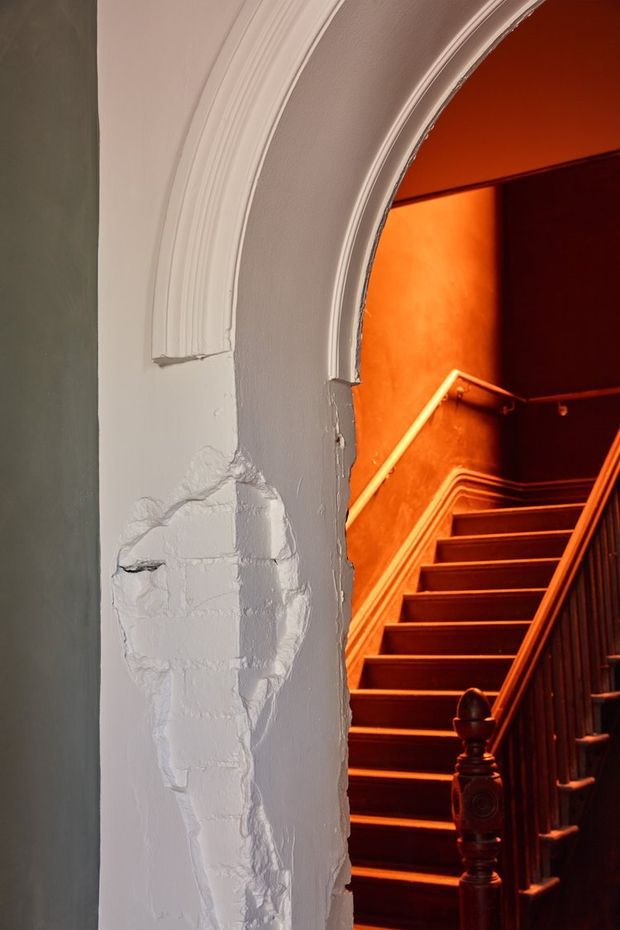 Close-up architectural interior view through a tall white plaster archway into a dramatically lit timber staircase. The arch has layered moulded trim and visibly weathered, patchy plaster, while the stair has dark stained timber treads, carved newel posts, balustrades and warm orange-red lighting. The image emphasises heritage detailing, texture and contrast between the pale foreground wall and moody stair hall.