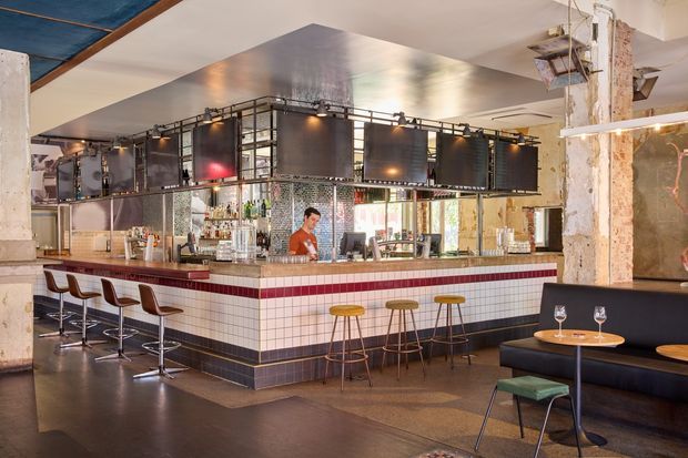 Interior view of an industrial-style commercial bar or restaurant. A large tiled bar counter with white square tiles, dark blue-grey lower tiles, and a red horizontal accent band is surrounded by stools and backed by a mirrored and glass bottle display. Dark metal-framed overhead shelving and a large metallic ceiling canopy dominate the space, while exposed, weathered structural columns and unfinished surfaces reinforce the industrial character. A banquette seating area is visible at right.