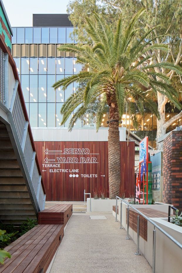 A landscaped urban commercial courtyard or access terrace with a palm tree, timber bench seating and decking, concrete-paved ramp, stainless-steel handrails, and external stairs. A red timber-clad wayfinding wall directs visitors to the Servo, Yard Bar, terrace, electric lane, and toilets. Behind it is a multi-storey contemporary building with extensive glazed curtain walls, dark horizontal cladding, and a patterned projecting facade screen. Brick walls, colorful signage, and mature trees frame the space.