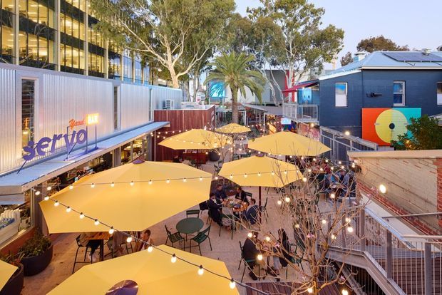 Elevated view of a lively urban outdoor dining precinct between contemporary commercial buildings. Multiple café tables sit beneath large yellow umbrellas, with festoon lighting strung overhead, stair access, murals, and landscaped trees creating a vibrant pedestrian courtyard atmosphere.