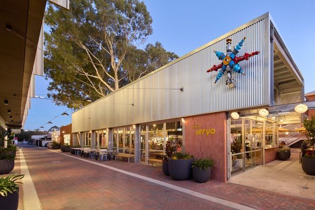 Contemporary low-rise commercial building viewed from a pedestrian plaza. The facade combines vertically ribbed light-metal cladding, extensive floor-to-ceiling glazing, and a brick-clad entrance volume. A colourful abstract wall sculpture provides a prominent feature, while outdoor seating, planters, and café-style lighting activate the paved public space. Mature trees and neighbouring buildings are visible in the background.