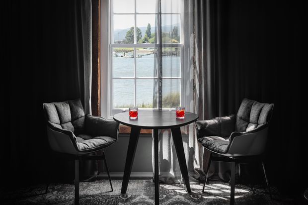 A compact, dramatically styled sitting or dining nook positioned in front of a tall multi-pane window. Two upholstered black lounge chairs flank a small round black table, with sheer curtains and heavy dark drapes framing the window. A patterned rug covers the floor, while a bright waterfront landscape with hills is visible beyond the glazing.