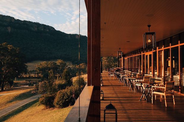 A long, covered timber deck or terrace beside a glazed building, furnished with rows of outdoor dining tables and chairs. Warm timber ceilings, decking, and framing create a contemporary hospitality setting, with expansive views toward a forested mountain or cliff landscape.