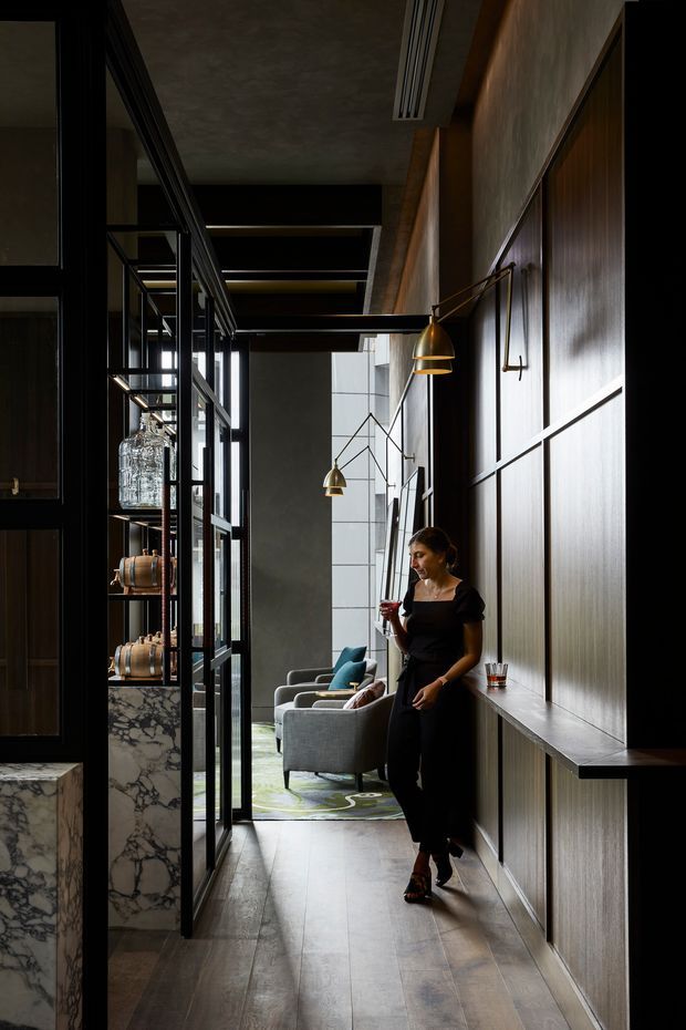 A dark, refined interior corridor or lounge approach with full-height black-framed glazed partitions and open shelving on the left, a marble-clad low cabinet, and a tall paneled timber feature wall with integrated console on the right. Sculptural brass wall lights illuminate the corridor, which leads to a seating area with grey armchairs, teal cushions, and a patterned rug. A person stands in the hallway holding a drink. The space has a sophisticated contemporary hospitality or residential entertaining feel, with strong vertical lines, layered lighting, and rich dark materials.