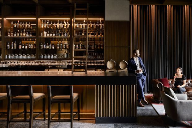 A sophisticated, dimly lit cocktail bar and lounge interior. The room features extensive backlit glass-fronted liquor shelving, a dark bar counter with upholstered stools, warm timber and fluted wall detailing, and a small seating area with armchairs and a sofa beside heavy dark curtains. The refined palette and layered lighting create an intimate hospitality atmosphere.