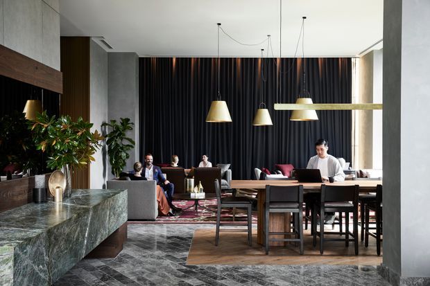 A sophisticated open-plan interior combining a dining area, lounge seating and a substantial stone island or counter. Dark floor-to-ceiling curtains, grey concrete-like walls and columns, richly veined stone, timber furniture, and sculptural pendant lighting create a refined, moody contemporary atmosphere. Indoor plants and occupants are visible, with daylight entering from the right.