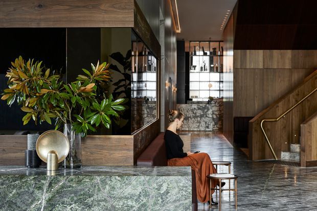 A sophisticated, dark-toned interior featuring a green marble reception or bar counter in the foreground, timber wall panelling and cabinetry, black-framed glazing, and a dramatic stairway with brass handrail. A seated person and indoor plants add life to the refined, hotel-like space. The rear area includes a marble-clad counter or feature wall and a large glazed opening, while linear ceiling lighting enhances the long circulation space.