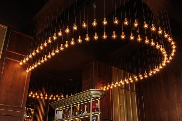 A dramatic, dimly lit interior featuring a large curved suspended installation of many warm glowing pendant bulbs above a bar. The space is clad in rich dark timber, with a sculptural layered bar canopy and open bottle shelving. The composition emphasizes the double-curved lighting feature and moody hospitality atmosphere.