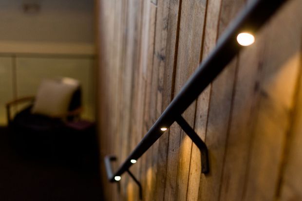 Close-up interior view of a rustic timber-clad wall with a black metal handrail and integrated small spotlights. A dimly lit seating area is softly visible in the background, creating a warm, intimate atmosphere.