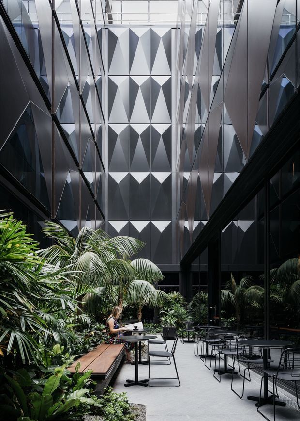 A dramatic, enclosed urban courtyard or atrium with lush tropical planting, outdoor café tables and chairs, reflective dark glazing, and tall faceted geometric metal wall panels rising through a multi-storey space. The composition emphasizes a contemporary commercial architectural interior/exterior hybrid with strong verticality, angular surfaces, and abundant greenery.