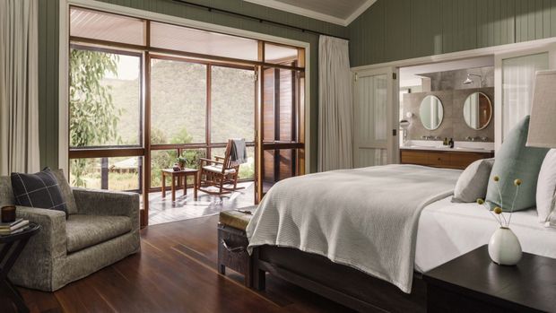 A spacious, refined bedroom with a dark timber floor, green vertical wall panelling, and a large glazed opening onto a furnished outdoor balcony or sunroom. The room contains a large upholstered bed with layered white and pale green bedding, bedside tables, an armchair, and a visible bathroom vanity through an open doorway. Warm timber window framing and abundant natural light create a relaxed, nature-connected atmosphere.