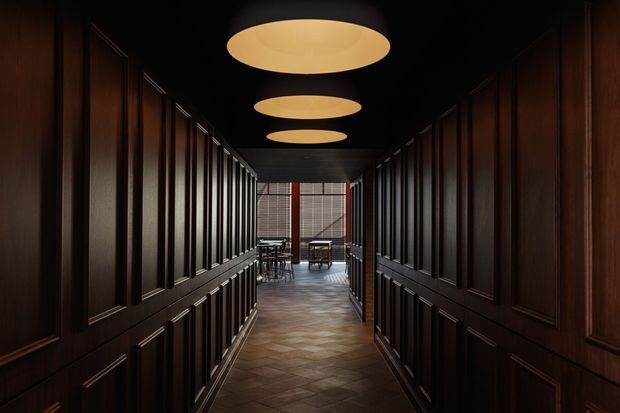 A dark, dramatic corridor lined with richly stained timber raised-panel wall panelling on both sides. The narrow hallway has a black ceiling with three large recessed circular pendant or flush-mounted ceiling lights, a patterned dark tile floor, and a dining or seating area visible at the far end beyond a glazed or screened opening.