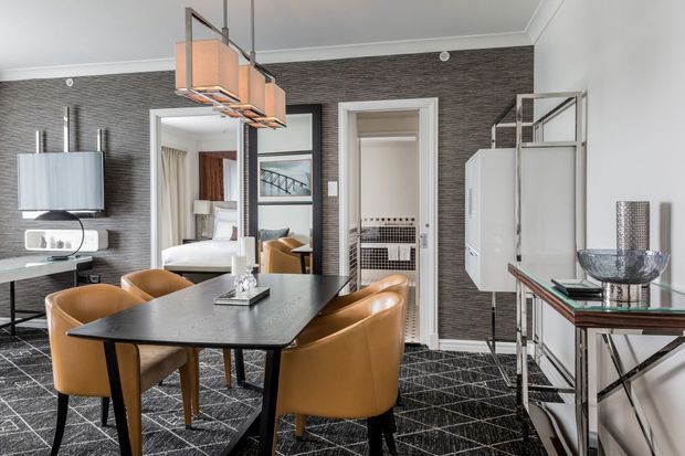 A refined dining room with a dark rectangular dining table surrounded by tan leather chairs. The room features a dark textured feature wall, geometric patterned carpet, contemporary pendant lighting, mirrored and glass furniture, and a sideboard with decorative objects. Open doorways reveal adjacent bedroom and bathroom areas.