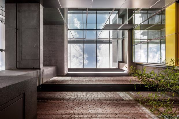 A dramatic modern architectural interior or semi-enclosed courtyard with exposed concrete walls, extensive black-framed glazing, steel structural elements, and a brick-paved floor. A glass bridge or upper-level walkway spans the bright double-height space, with reflected daylight and a small planted area adding contrast.