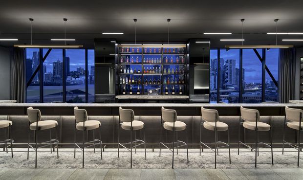 A sophisticated contemporary home bar or hospitality-style bar interior with a long dark counter and a row of cream upholstered bar stools. The back bar features illuminated glass-front shelving filled with bottles, flanked by large windows overlooking a dense city skyline and waterfront at dusk. Dark ceilings, integrated linear lighting, black structural framing, and polished dark finishes create a dramatic, refined atmosphere.