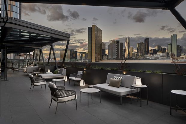 A sophisticated rooftop terrace overlooking a dense modern city skyline at dusk. The expansive outdoor space features dark tiled flooring, black steel framing and screening, contemporary lounge seating, small round tables, planters, and a covered dining or lounge zone. The atmosphere is refined and urban, with warm interior-like lighting contrasting against the cool evening sky.