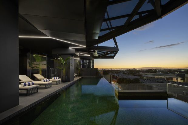A dramatic rooftop pool terrace at dusk, featuring a long infinity-edge swimming pool beside a dark, contemporary pavilion. The terrace includes cushioned loungers, large potted plants, integrated linear lighting, and a striking glazed and steel cantilevered roof structure. The pool overlooks a low-rise urban landscape and distant horizon.