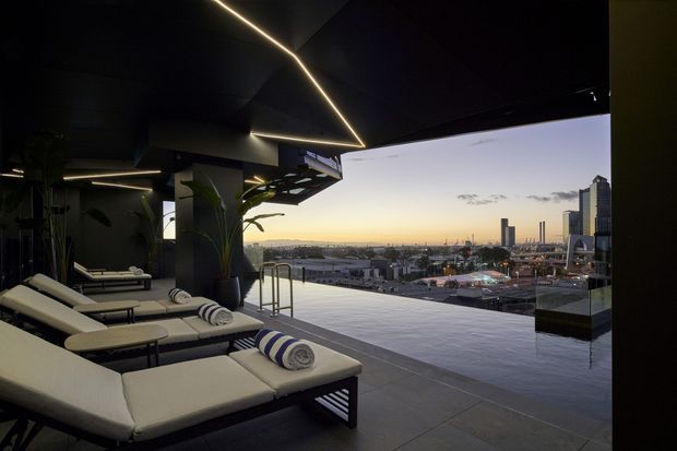 A sophisticated rooftop pool terrace at dusk, with a dark sculptural canopy and linear integrated lighting over several cream upholstered sun loungers. A narrow infinity-edge pool and glass balustrade overlook a dense urban skyline, while large potted plants soften the otherwise minimal setting.