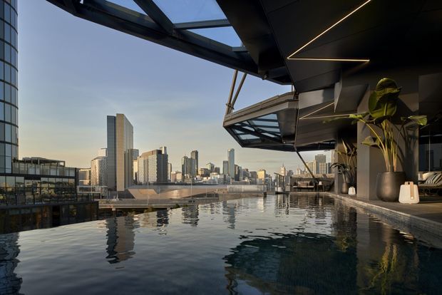 A rooftop infinity pool or water feature overlooks a dense modern city skyline. The scene is viewed from a sophisticated contemporary rooftop terrace with dark metal soffits, angular projecting balconies, extensive glazing, integrated linear lighting, and large potted plants. Calm reflective water dominates the foreground, while high-rise buildings and a waterfront-looking urban setting extend beyond.