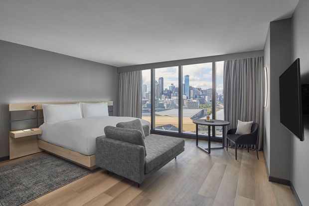 A contemporary hotel-style bedroom with a neatly made bed, upholstered grey lounge chair and ottoman, small round side table, bedside furniture, wall-mounted television, and full-height curtains framing expansive city views. The room has grey walls, light timber flooring, and a calm, refined neutral palette.