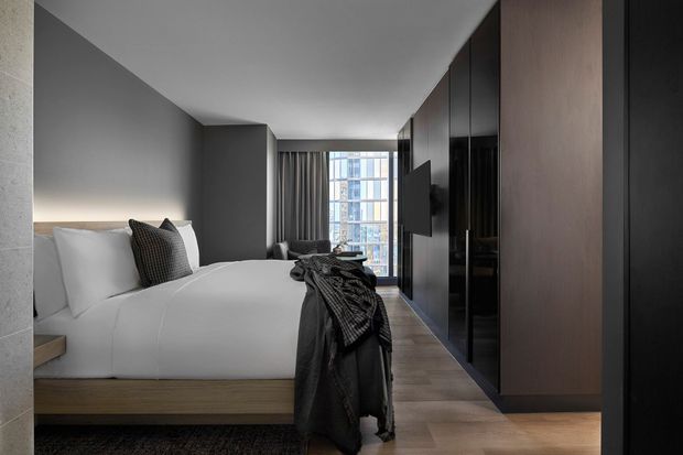 Contemporary bedroom interior with a low timber bed, crisp white bedding, charcoal cushions and throw, and a dark floor-to-ceiling built-in wardrobe and media wall. Grey walls, timber flooring, full-height curtains, and a large glazed window create a refined, moody urban atmosphere.