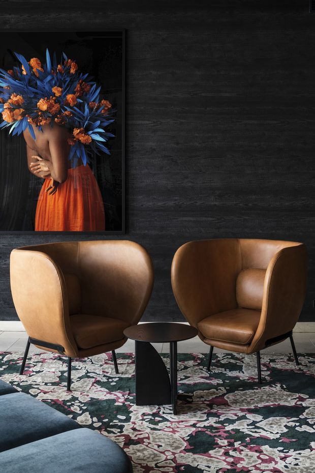 A moody, refined seating area with two caramel-brown leather wingback lounge chairs arranged around a small dark round coffee table. A richly patterned floral rug covers the floor, while a dark textured wall with framed floral artwork forms the backdrop. The corner of a deep blue sofa is visible in the foreground.