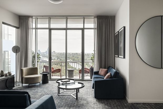 A refined contemporary living room with floor-to-ceiling glazed doors opening to a balcony, overlooking a leafy urban landscape. The room features a dark blue sofa, sculptural lounge chairs, round glass-and-metal coffee tables, full-height grey curtains, grey patterned carpet, minimalist framed wall art, and a large round wall mirror.