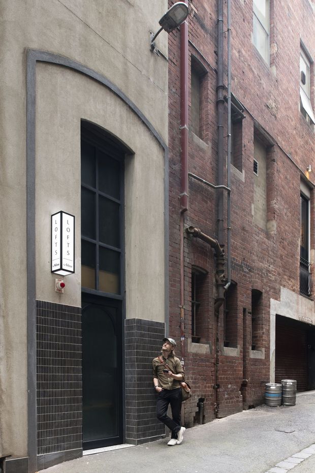 A narrow urban laneway showing the exterior of a converted brick building. The facade combines aged red brickwork, rendered concrete, arched openings, dark metal-framed glazing, exposed drainage pipes, and a tiled lower wall section. A projecting sign identifies an office entrance, with a person standing beside the doorway and metal barrels near a garage-like opening.