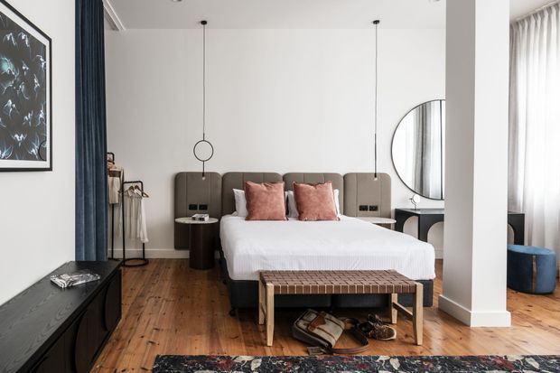 Contemporary bedroom with a low upholstered bed and dark taupe headboard, white bedding, blush pink cushions, and a timber slat bench at the foot. The room features warm timber flooring, white walls, floor-length curtains, pendant lights, a round wall mirror, and dark built-in or freestanding cabinetry. A partial adjoining seating area is visible beyond the central column.