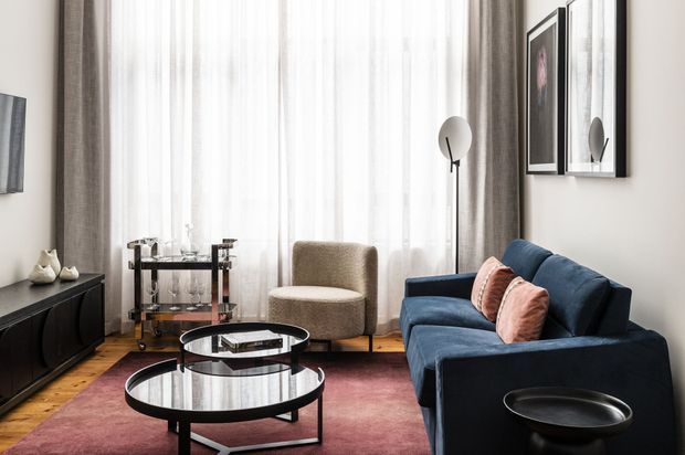 Refined contemporary living room with a tall sheer-curtained window, dark blue sofa, rounded boucle-style armchair, nested circular glass coffee tables, black media console, floor lamp, framed artwork, and a muted burgundy rug over timber flooring.