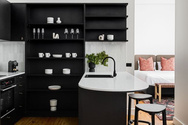 A compact, open-plan kitchen with a black built-in shelving wall, dark cabinetry, white stone island benchtop, and integrated sink with curved black tap. White tableware and glassware are displayed on the shelving, while a bedroom area with blush cushions is visible behind the island. Timber flooring, a patterned rug, and black stools add contrast to the monochrome fit-out.