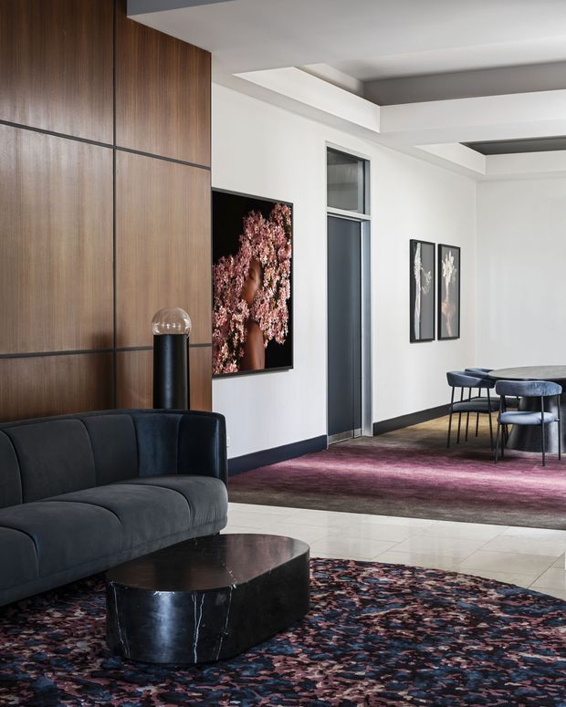 A refined contemporary lounge and dining area with a dark charcoal sectional sofa, sculptural black coffee table, patterned area rugs, walnut wall panelling, black-framed internal glazing, and a compact dining setting. White walls and a layered recessed ceiling provide contrast, while burgundy and mauve tones in the rugs add depth.