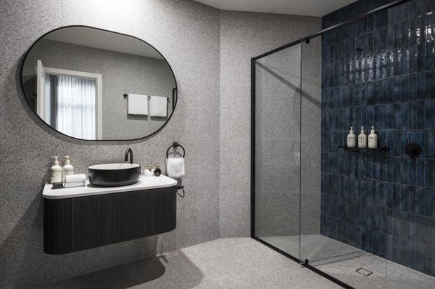 Contemporary bathroom featuring a floating dark ribbed vanity with a black vessel basin, large rounded-edge mirror, frameless glass shower enclosure, grey textured wall and floor finishes, and a deep blue tiled shower feature wall.