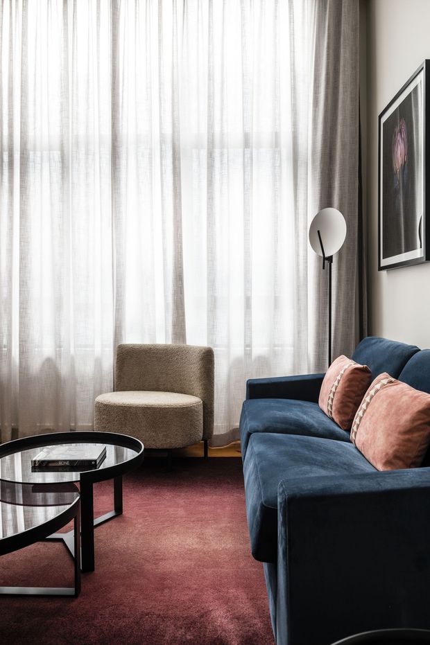 A refined living room with a deep blue velvet sofa, blush pink cushions, a rounded boucle armchair, black-and-glass coffee table, burgundy carpet, sheer floor-to-ceiling curtains, and a sculptural globe floor lamp. Framed artwork and a neutral wall complete the sophisticated, high-contrast interior.