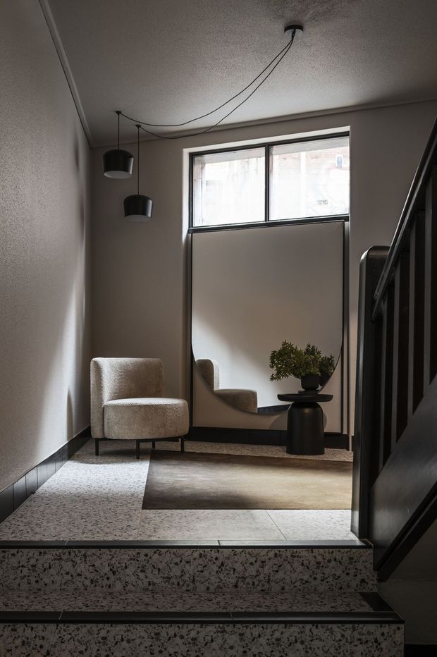 A refined residential stair landing with a small upholstered lounge chair, round black pedestal table, potted plant, wall mirror, and two suspended pendant lights. The space has dark timber stair and trim elements, pale textured walls, a black-framed window, terrazzo stair treads and landing, and a muted neutral rug. The atmosphere is calm, moody, and minimalist with warm contemporary detailing.