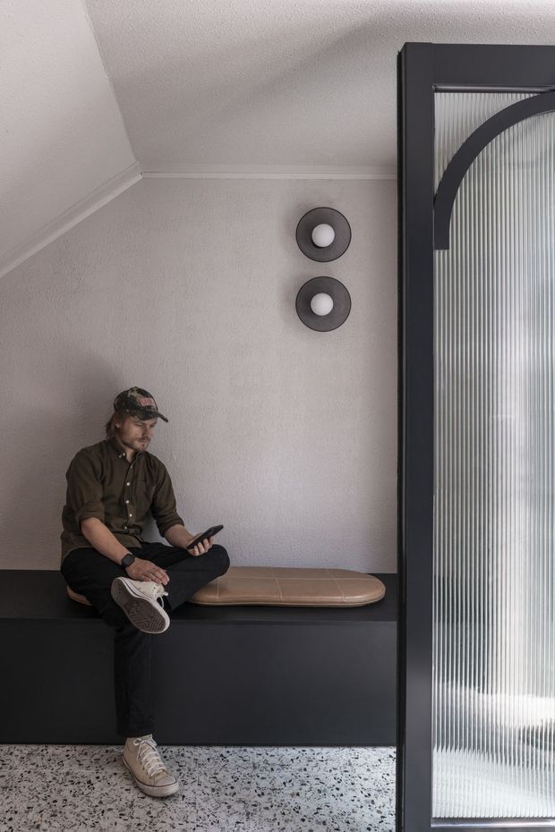 A compact contemporary interior nook with a built-in dark charcoal bench, tan upholstered seat cushion, textured light plaster walls, and a speckled terrazzo floor. A person sits on the bench beside a tall dark-framed ribbed translucent glass door or screen. Two circular wall lights add subtle decorative detail.