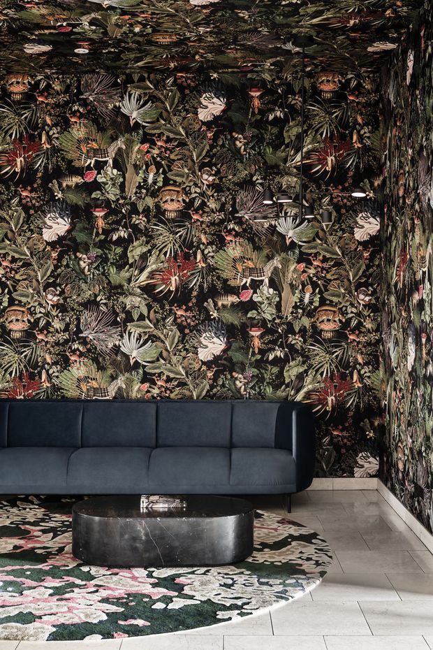 A dramatic living room featuring floor-to-ceiling dark botanical wallpaper on the walls and ceiling, a deep navy upholstered sofa, a round black marble coffee table, and a large floral-patterned area rug over pale tiled flooring. Minimal black pendant lighting and strong patterned surfaces create a bold, luxurious atmosphere.