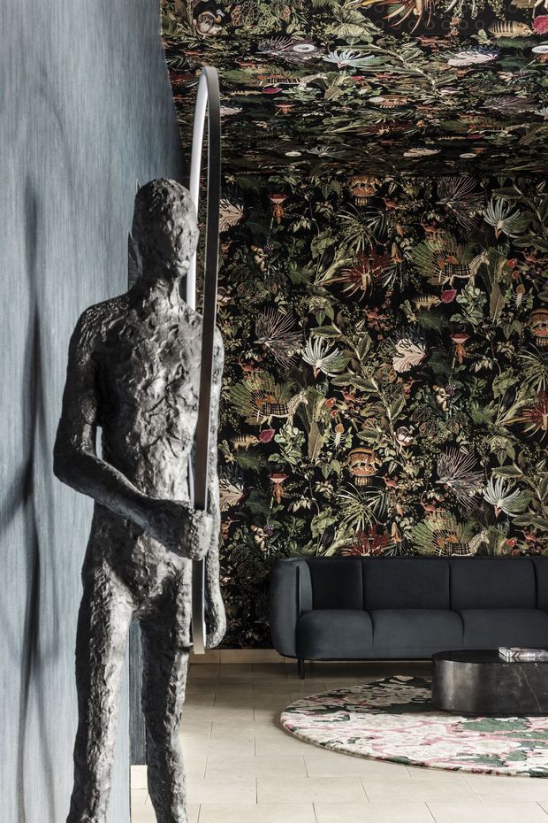 Dramatic, moody interior featuring a dark botanical patterned wallcovering extending across the wall and ceiling, a charcoal upholstered sofa, pale tiled flooring, and a sculptural metallic figure holding a tall oval form in the foreground. A patterned round rug and low dark coffee table complete the richly layered, art-forward lounge setting.