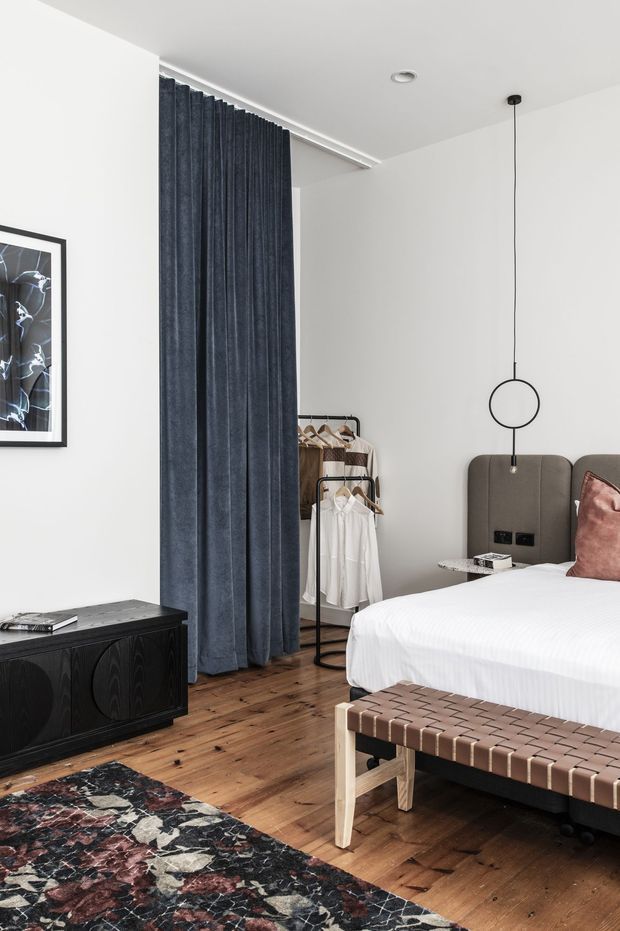 A contemporary bedroom with white walls and ceiling, warm timber flooring, a dark blue full-height curtain concealing an open wardrobe area, and a bed with a tall upholstered headboard. A black timber sideboard, woven leather bench, patterned rug, framed artwork, and minimalist pendant light complete the refined interior.