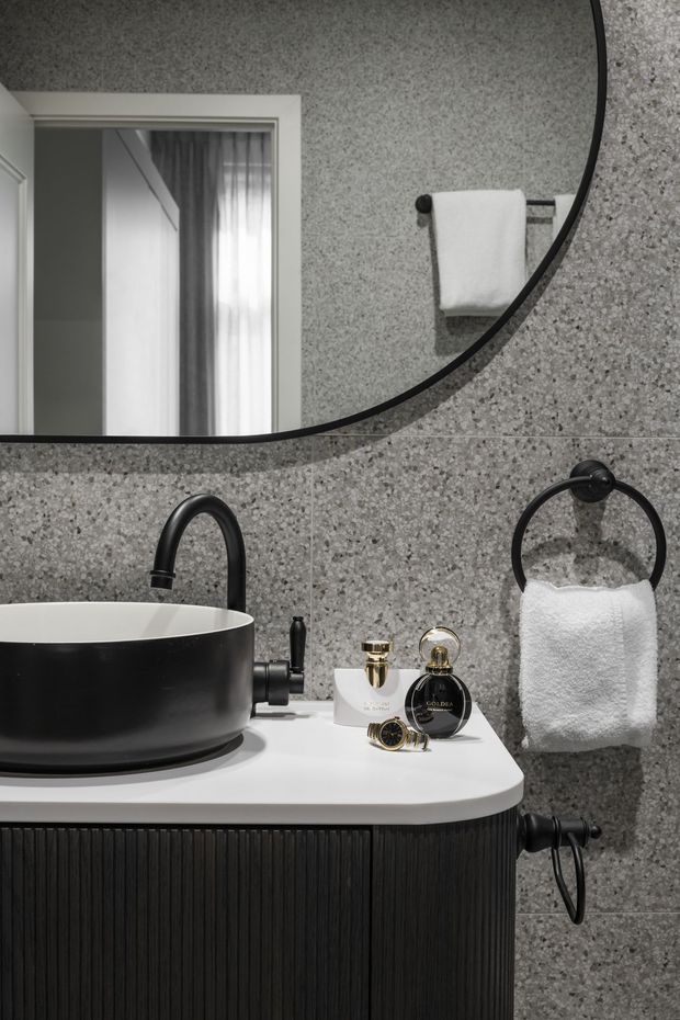 Close-up view of a refined bathroom vanity featuring a round black vessel basin, matte black tapware, a white solid-surface countertop, and dark fluted cabinetry. The walls are clad in grey terrazzo-like stone tiles, with a large round black-framed mirror and matching circular towel rings. Toiletries sit on the benchtop, while a doorway and curtains are reflected in the mirror.
