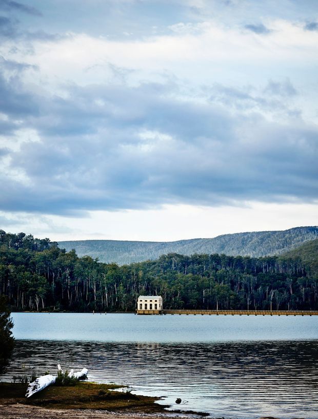 A tranquil landscape photograph of a lake surrounded by dense forest and wooded mountains under a dramatic cloudy sky. A small pale building stands at the end of a long narrow pier across the water. The foreground includes a low vegetated shoreline and a bird, while the scene is dominated by the natural waterfront setting rather than a visible architectural interior.