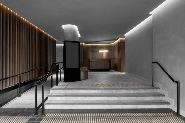 A refined, dimly lit architectural entry or circulation space with broad stone steps, black metal handrails, dark tiled flooring, and a dramatic slatted timber feature wall illuminated by linear lighting. The stairway leads to a recessed lobby with curved wood-toned wall panels, integrated lighting, and a sculptural light feature. The overall design is minimalist and contemporary with a restrained dark neutral palette.