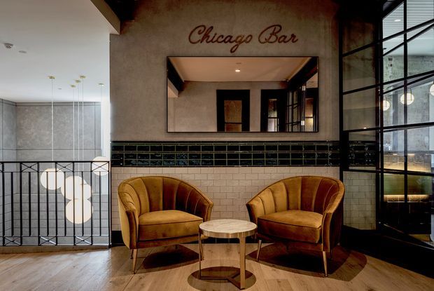 A stylish Chicago-themed bar lounge with two mustard leather tub chairs and a small round table in front of a tiled feature wall. The wall includes a large mirror, black-and-green decorative tile band, white subway tile, and painted script signage. Black metal-framed glazing and railings, pendant lighting, and warm timber flooring create a refined industrial Art Deco-inspired atmosphere.