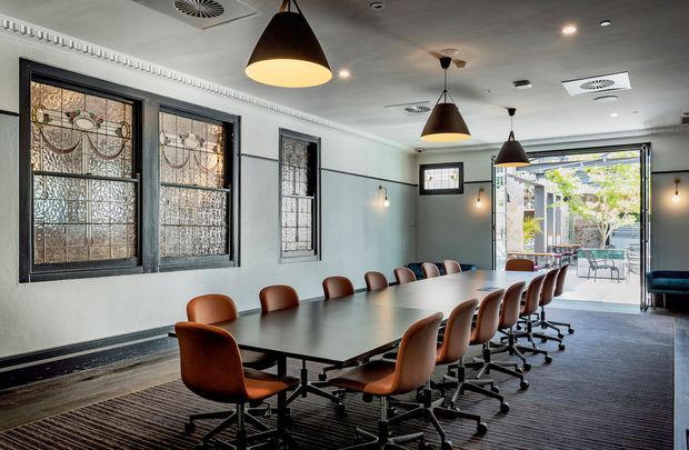 A contemporary meeting room with a long dark conference table surrounded by tan leather swivel chairs. The room features decorative stained-glass windows in dark frames, pale walls with detailed cornice moulding, dark carpet, pendant lighting, and glazed doors opening toward an outdoor courtyard or terrace.