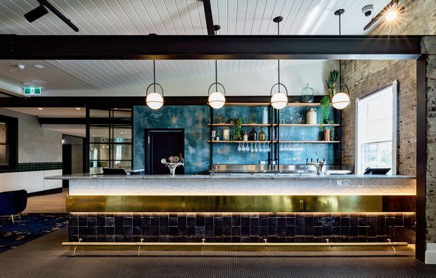 A stylish commercial bar or hospitality counter with a long stone-clad benchtop, dark glossy tiled front, illuminated brass-toned kickboard, open timber shelving, and a blue textured feature wall. The space combines exposed masonry, black steel framing, white linear ceiling panels, globe pendant lights, and contemporary industrial detailing.