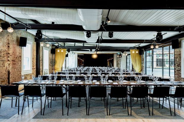 A large industrial-style event dining room arranged for a formal function, with long rows of tables set with white place settings and glassware, black chairs, exposed brick walls, large factory-style windows, visible ductwork, black structural beams, and theatrical lighting. Gold curtains frame the rear opening and add a warm accent to the otherwise neutral space.
